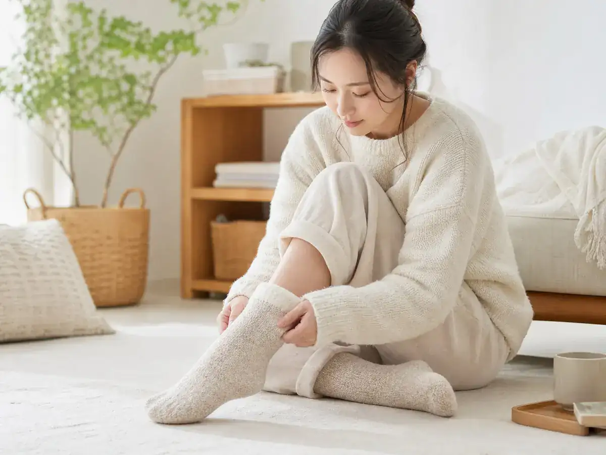 ニットの部屋着姿の女性が、床に座ってウールの靴下をはいている写真